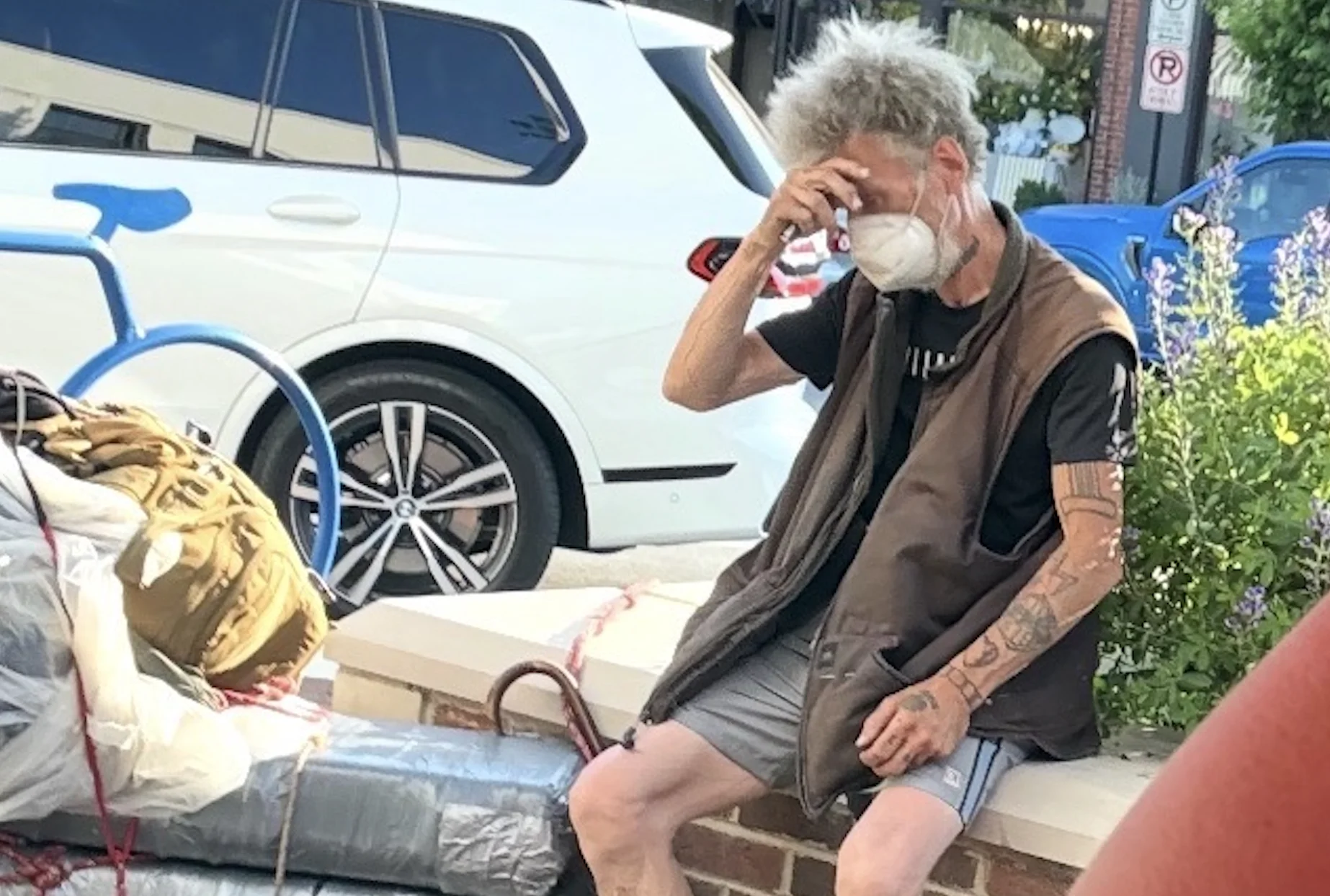 Homeless man sits beside a bundled pile of belongings next to a planter in downtown Glen Ellyn, underscoring ongoing concerns among some residents about visible homelessness in public spaces.